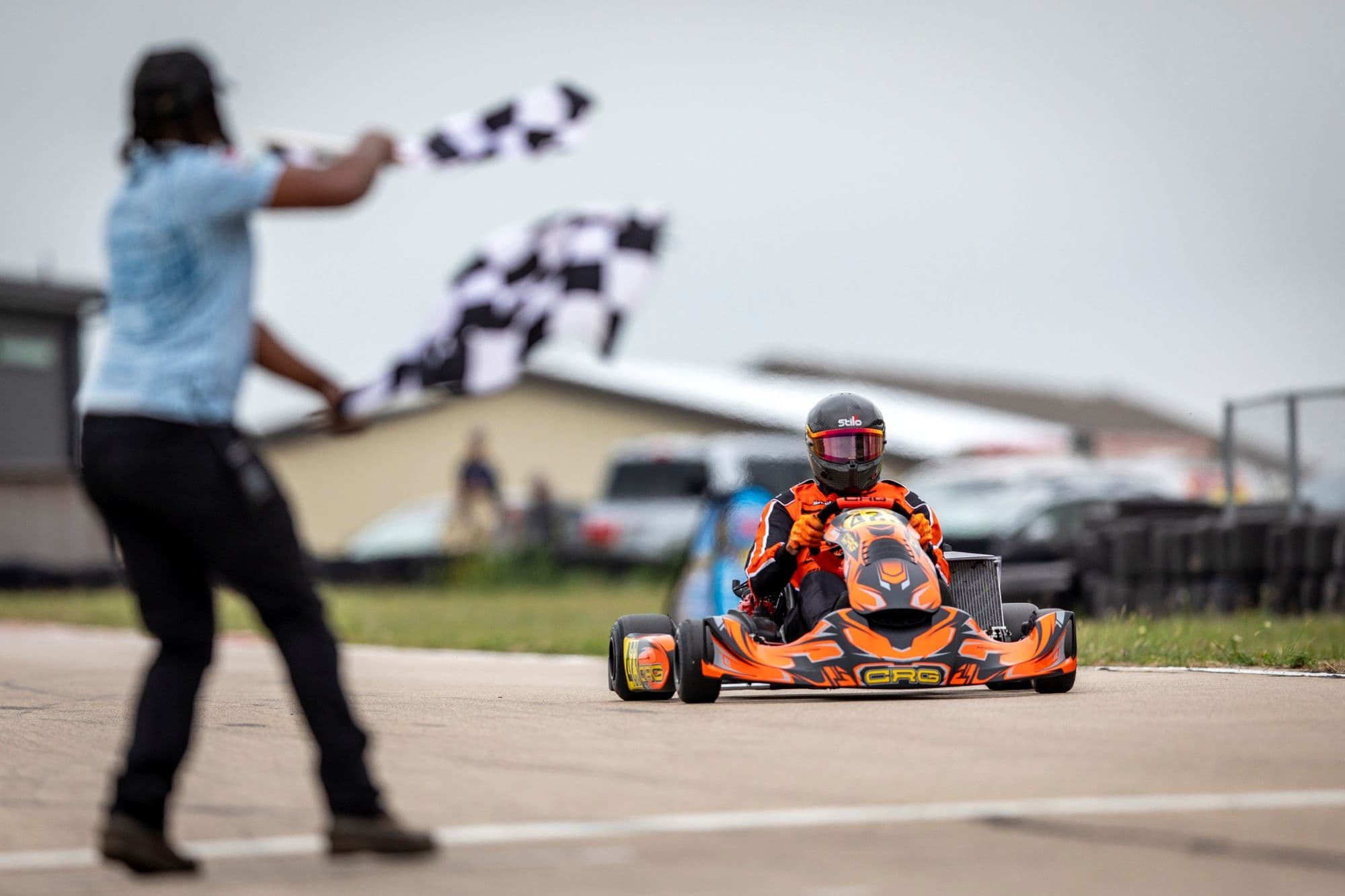 A flagman waves the checkered flag as an orange kart takes the finish at KartMoto.