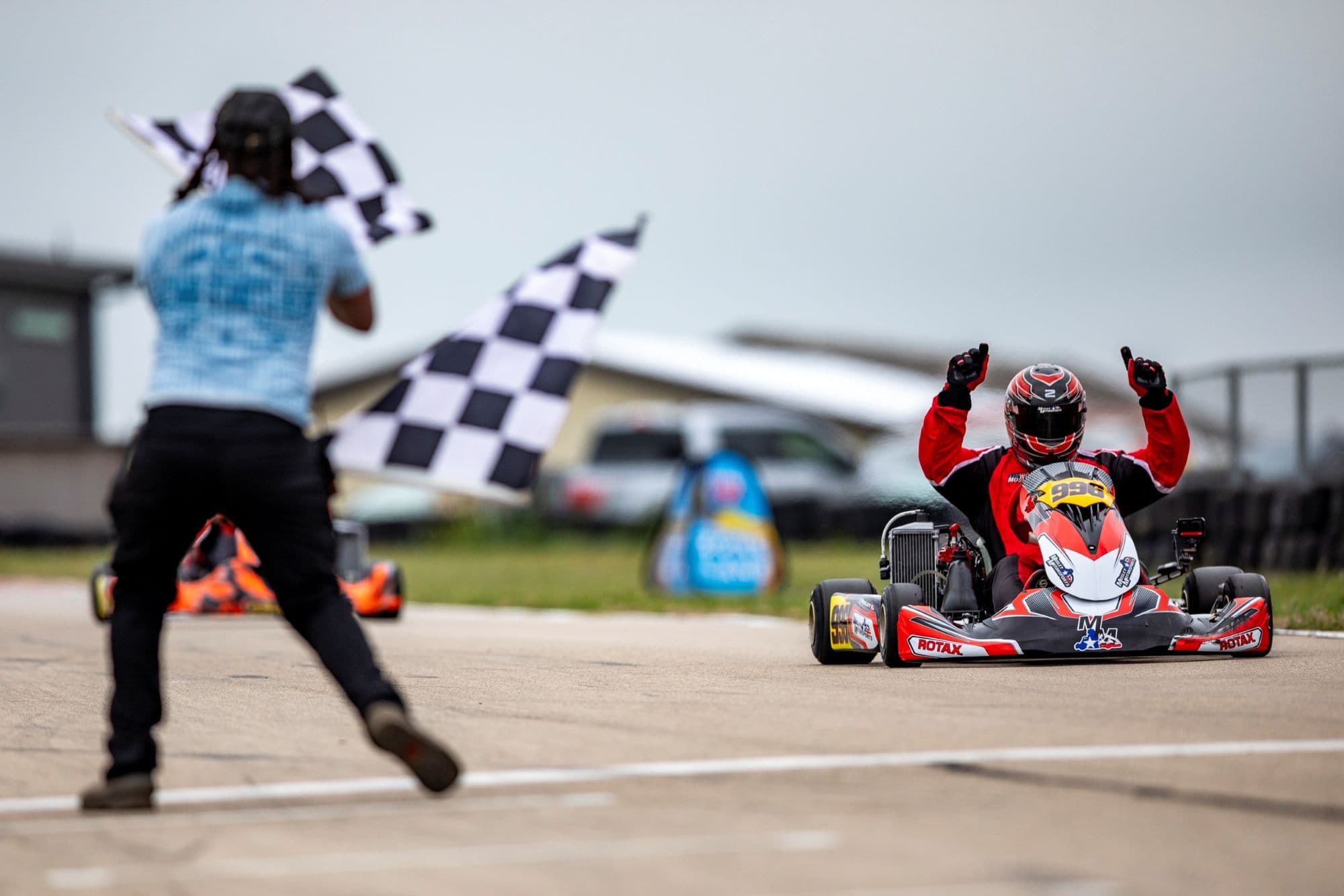 The checkered flag waves while a driver celebrates with both arms raised at KartMoto.