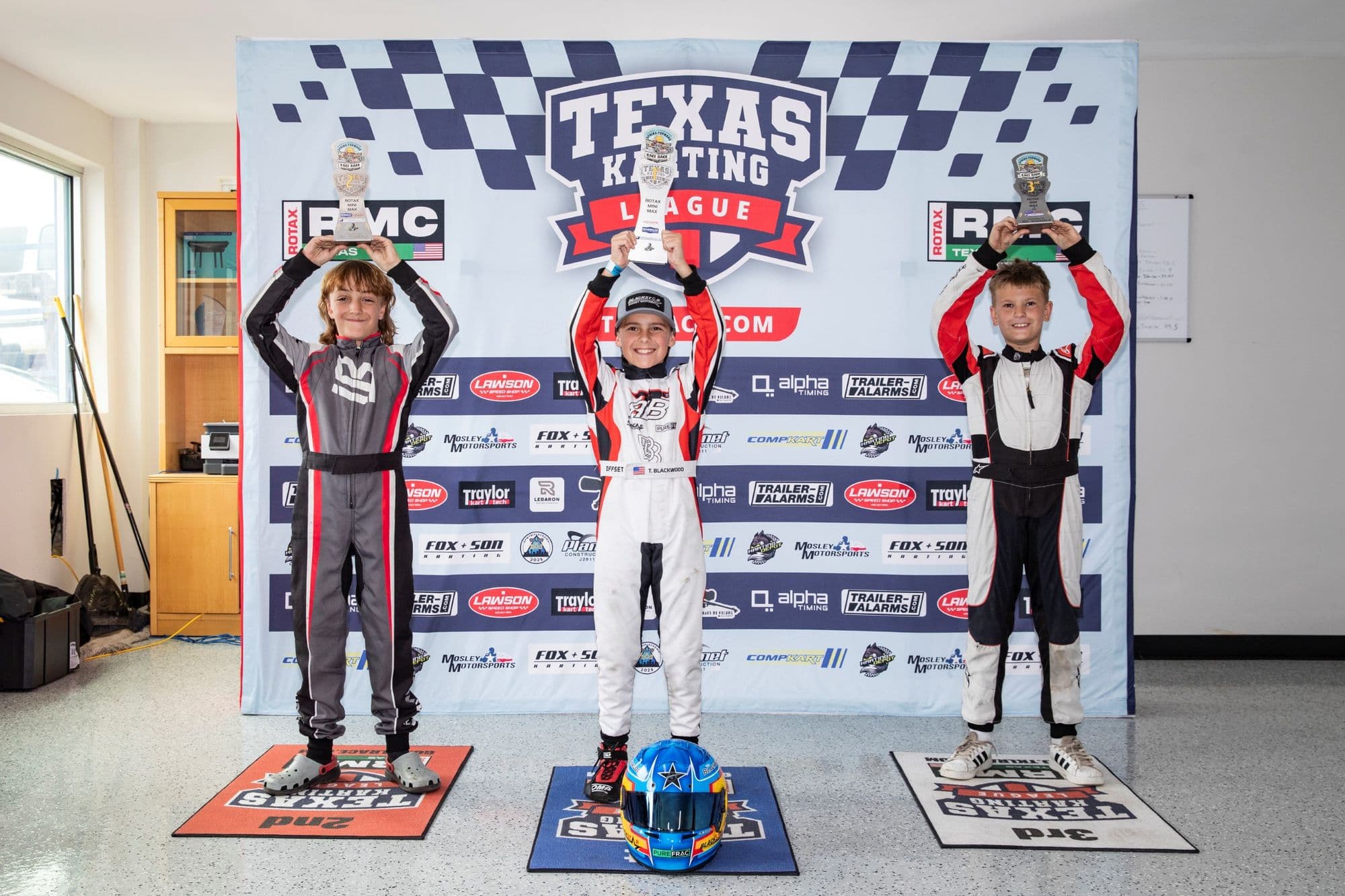 Three drivers in gray and black suits raise trophies on the KartMoto podium.