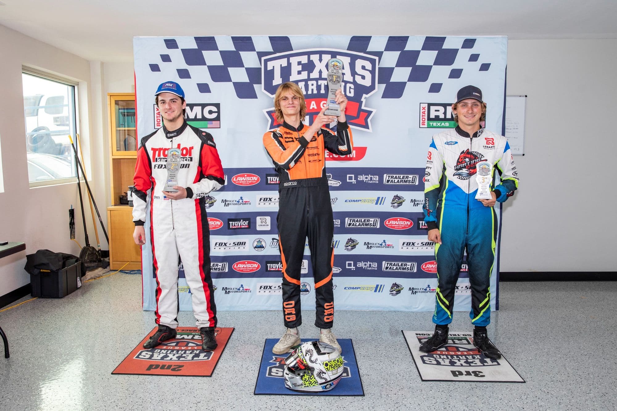 Three drivers in colorful suits stand with trophies at KartMoto.